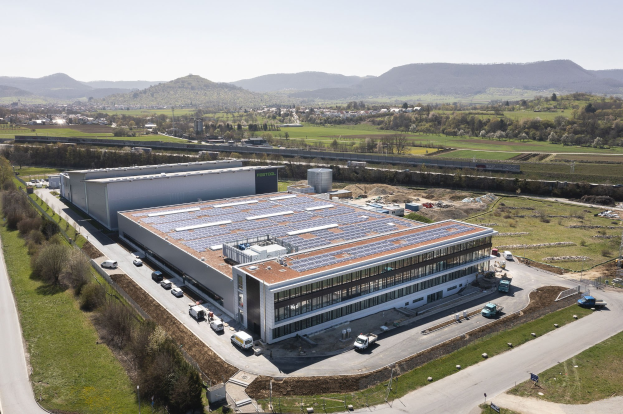 Aerial view of a large building with solar panels on its roof, surrounded by vehicles, trees, grass, and poles, with hills and a clear blue sky in the background; the building is Germany's first solar-powered factory.
