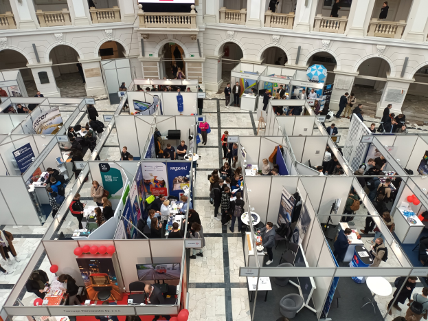 Große Gruppe von Menschen auf einer internationalen Messe mit Ständen, Bannern, Tischen, Stühlen und Ballons in einem Raum mit architektonischen Bögen und Säulen.