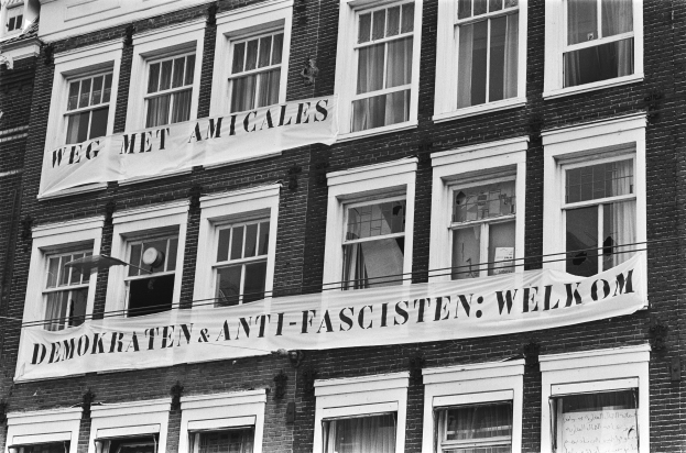 Black and white photo of a building with windows and a banner reading "Demokraten & Anti-Fascisten Welkom" attached to the wall.