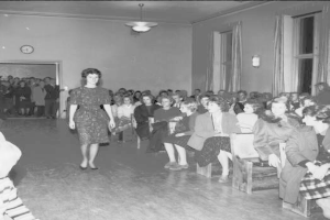 Eine Frau in einem schwarzen und weißen Kleid läuft über einen Laufsteg während einer Modenschau für Frauenrechte, mit einer Menge, die auf Stühlen sitzt und einer Wand mit Uhr und Beleuchtung im Hintergrund.