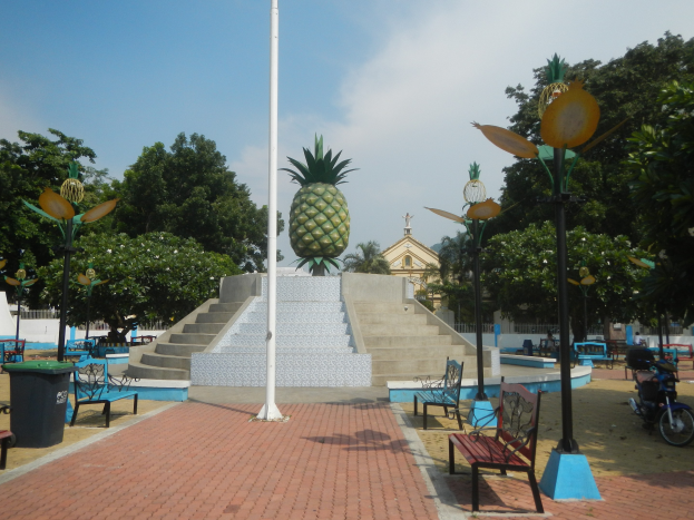 A park scene featuring benches, tables, trash bins, light poles, lampposts, stairs, a pineapple statue, a building, trees, and a cloudy sky.