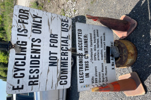 Ein Schild mit der Aufschrift "E-Bike-Fahren ist nur für Anwohner" steht an der Seite einer Straße, umgeben von Verkehrskegeln und anderen Gegenständen, mit einem Fahrzeug, Bäumen und einem klaren blauen Himmel im Hintergrund.