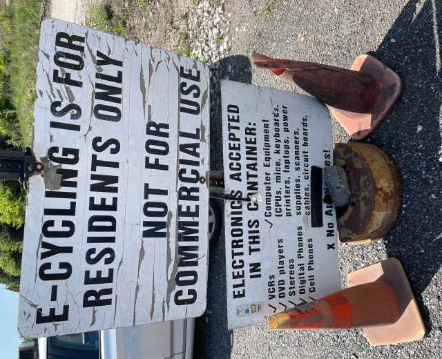 Ein Schild mit der Aufschrift "E-Bike-Fahren ist nur für Anwohner" steht an der Seite einer Straße, umgeben von Verkehrskegeln und anderen Gegenständen, mit einem Fahrzeug, Bäumen und einem klaren blauen Himmel im Hintergrund.