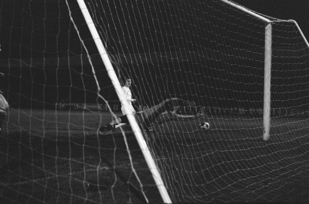 Ein Fußballer in einem Schwarz-Weiß-Foto hält den Ball, mit einem Ball in der Luft und einem Tor im Hintergrund.