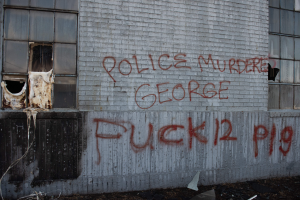 Ein Gebäude mit Graffiti, das 'Polizei Mord George Fuck 12 P19' auf der Seite trägt, mit Fenstern und diversen Gegenständen davor.