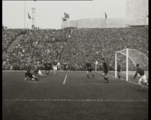 Schwarzes und weißes Foto vom WM-Finale 1958 zwischen Manchester United und Liverpool, das Spieler auf dem Feld, ein Torpfosten auf der rechten Seite, Zuschauer in den Rängen und Fahnen im Hintergrund zeigt.
