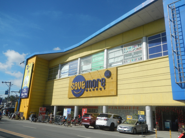 Ein gelbes und blaues Geb├Ąude mit einer "Mehr sparen"-Aufschrift, umgeben von Fahrzeugen, Passanten, Strommasten, B├Ąumen und einem bew├Âlktem Himmel.