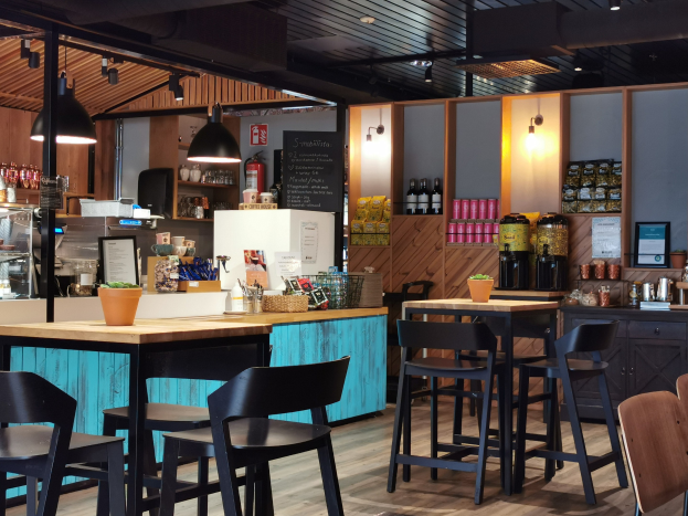 Innenraum eines Restaurants mit Holztischen, -stühlen, einer Topfpflanze auf einem Tisch, einer Schiefertafelmenü, einem Kühlschrank, einem Feuerlöscher, Regalen mit Flaschen, Schränken mit Gegenständen, einem Rahmenfoto an der Wand und Deckenleuchten.