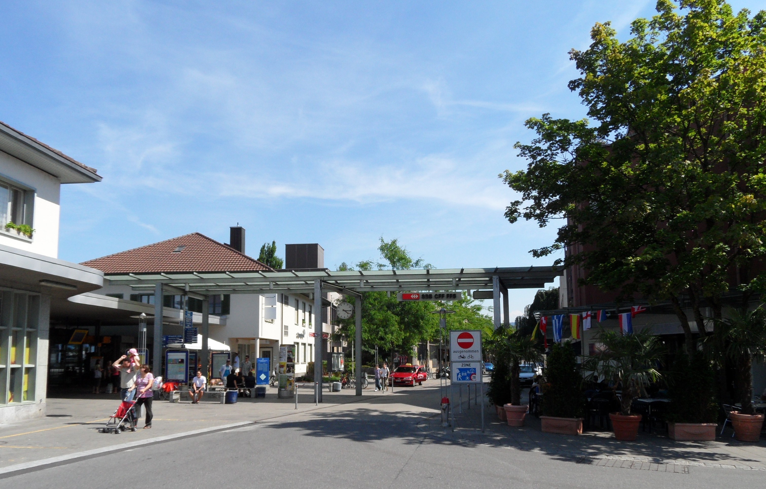 Stadtstraße mit Fußgängern, darunter einige mit Kinderwagen, gesäumt von Gebäuden, Bäumen, Topfpflanzen, Schildern und Fahrzeugen unter einem bewölkten Himmel.