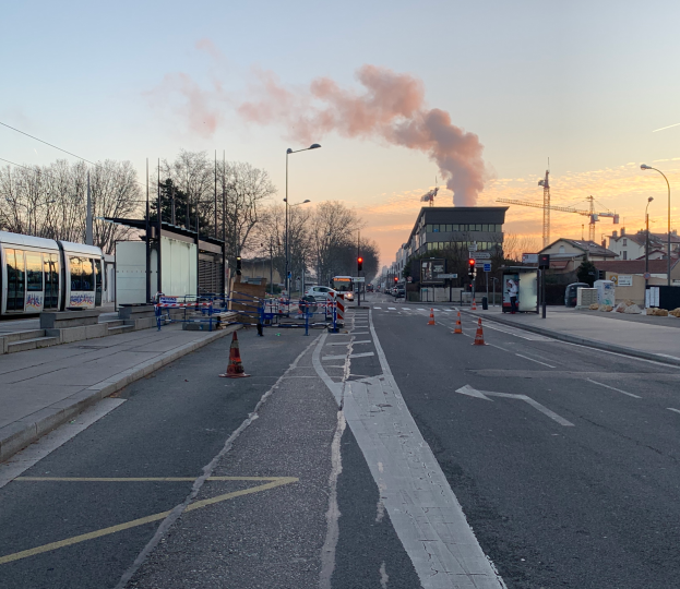 Eine Straßenkreuzung mit Verkehrskegeln, einem Bus, Laternen, Bäumen, Gebäuden, Kränen und Rauch, der aus einem fernen Gebäude aufsteigt.