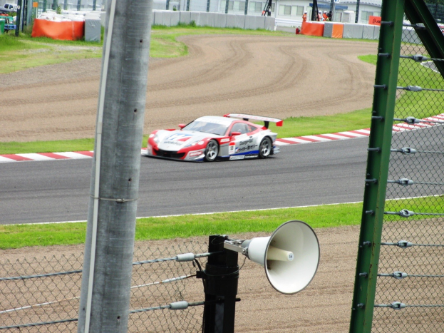 Ein Rennwagen fährt auf einer Rennstrecke mit einem Megafon vorne daran, umgeben von einem Metallzaun, Gras, Gebäuden, Bäumen und einem klaren blauen Himmel.