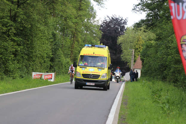 Ambulanz fährt auf einer Straße mit Fahrradfahrern daneben, Gras und Bäume auf beiden Seiten, Häuser, Pfähle und einen klaren blauen Himmel im Hintergrund.