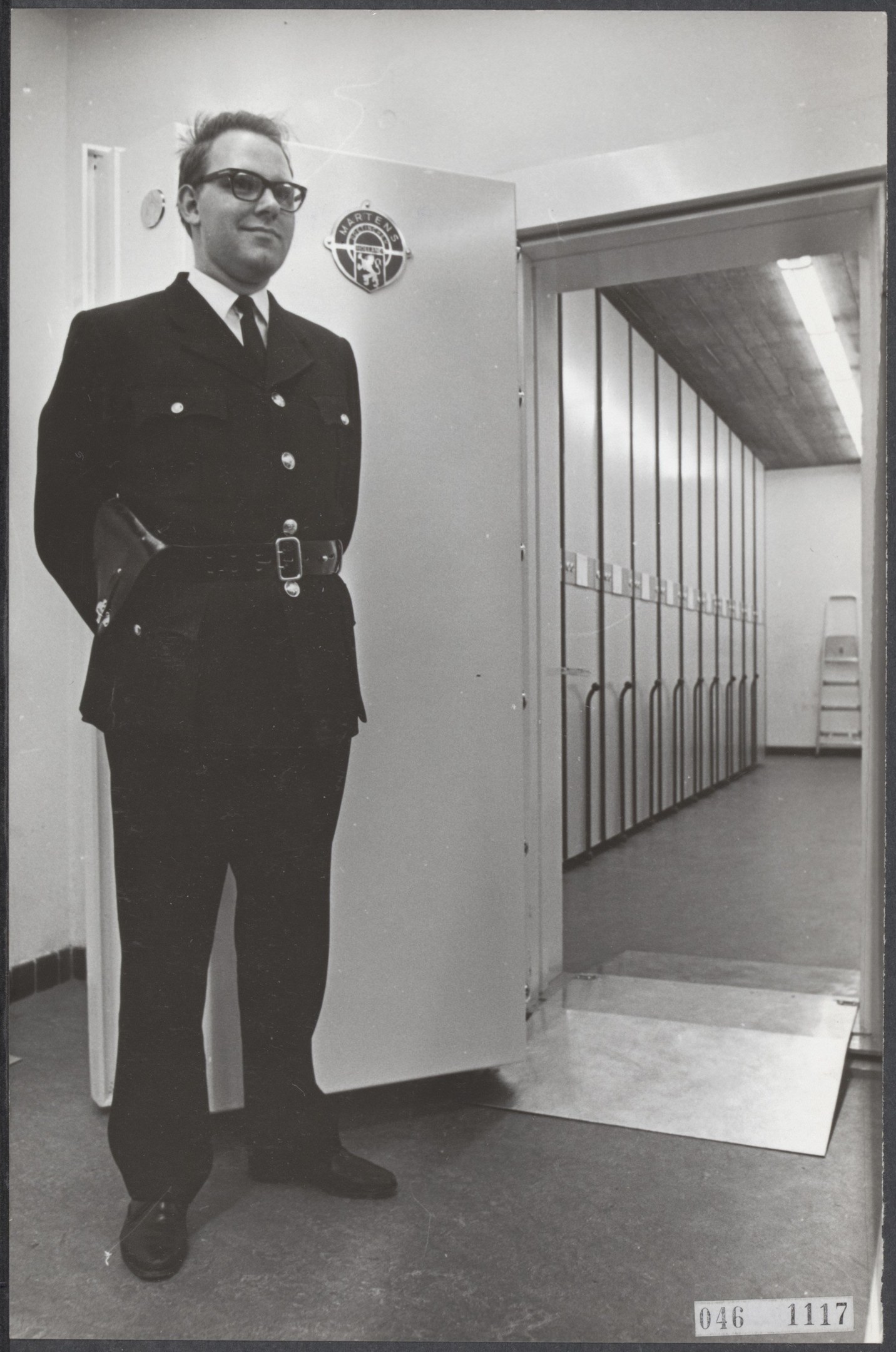 A police officer in uniform stands in front of lockers, wearing a coat, tie, shirt, trousers, shoes, and spectacles, with a wall, door, ladder, and ceiling in the background.