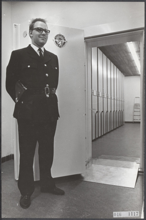 A police officer in uniform stands in front of lockers, wearing a coat, tie, shirt, trousers, shoes, and spectacles, with a wall, door, ladder, and ceiling in the background.
