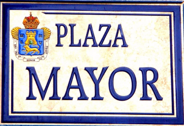 Ein rechteckiges weißes Schild mit schwarzer Schrift, die 'Plaza Mayor' lautet und einen blauen Rahmen hat, mit einem gekrönten Wappenlogo darauf, das an einem Gebäude angebracht ist.