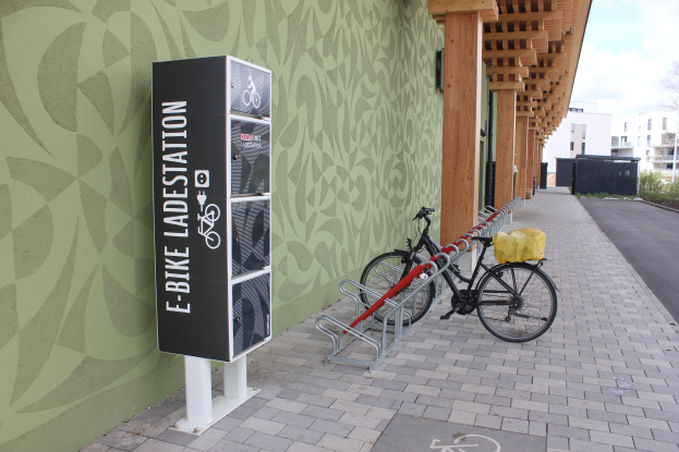 Ein Fahrrad, das vor einem Gebäude mit einem "E-Bike-Ladestation"-Schild geparkt ist, mit einem Fahrradrad im Vordergrund, Gebäuden, Bäumen, Pflanzen und einem klaren blauen Himmel im Hintergrund.