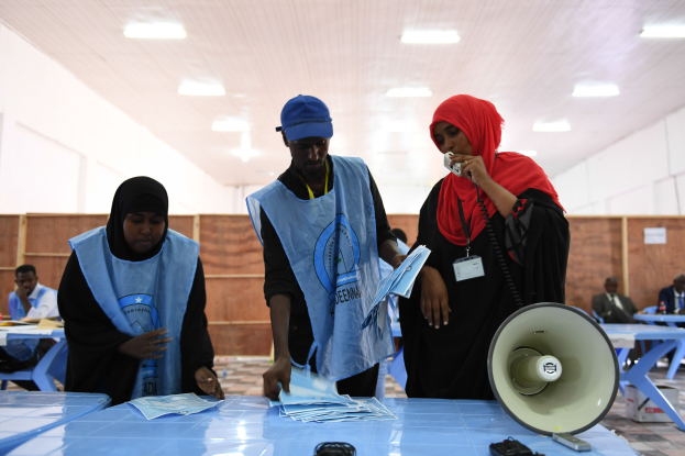 Menschen versammeln sich um einen Tisch mit Megafon und Papieren an einer Wahllokal, während andere im Hintergrund auf Stühlen sitzen und stehen, unter beleuchteter Decke.