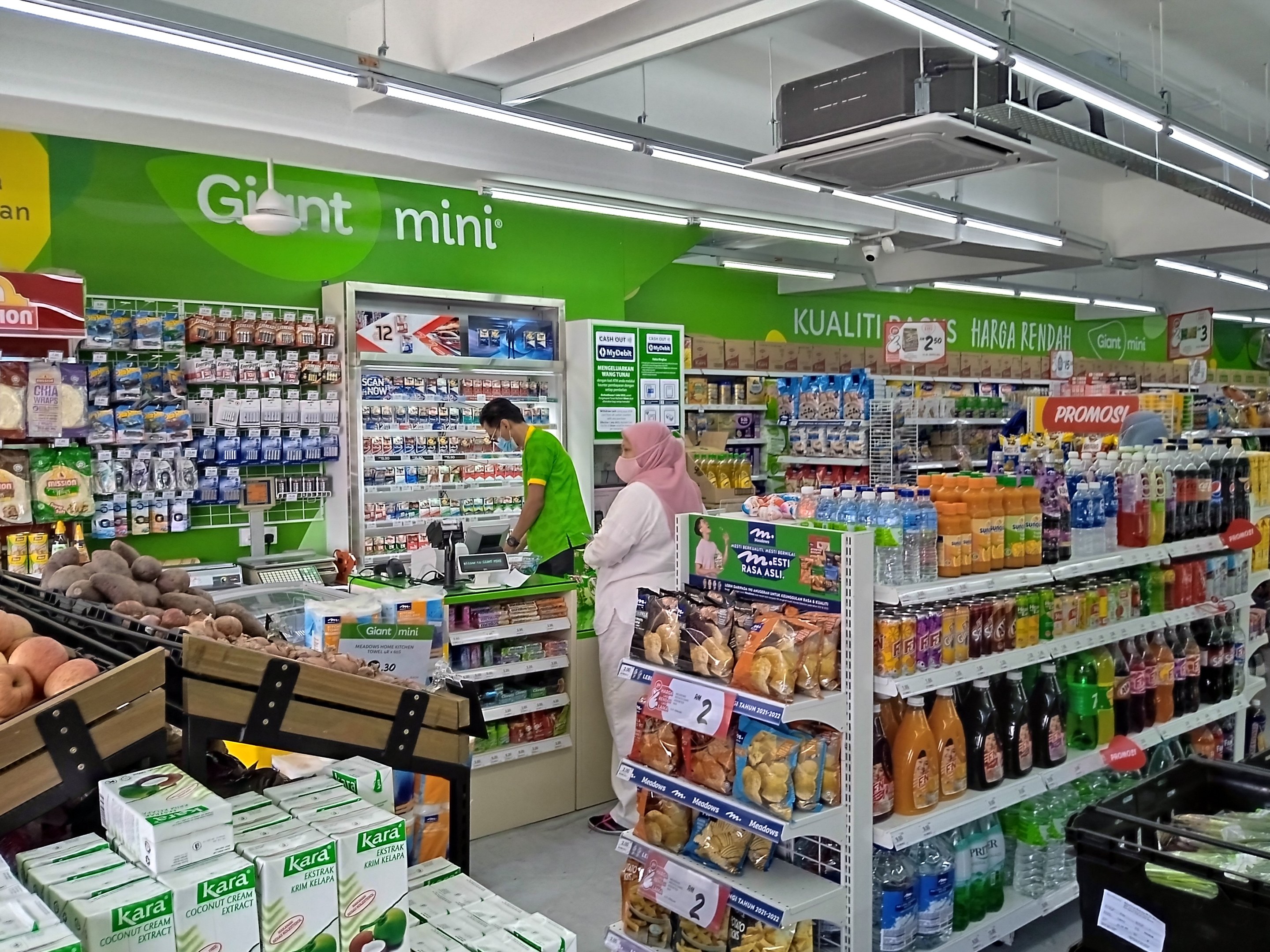 A brightly lit mini supermarket aisle stocked with bottles, boxes, fruits, and packaged goods on shelves, featuring ceiling-mounted lights, text boards, and a projector.