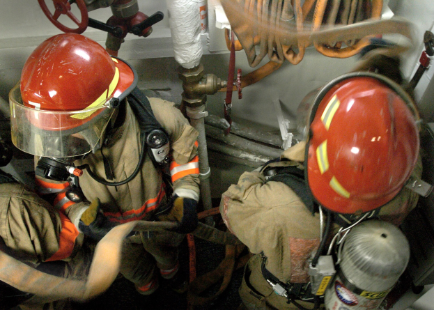 Feuerwehrleute in Schutzausrüstung bei der Arbeit an einem Feuerhydranten mit Leitungen und Ventilen im Hintergrund.