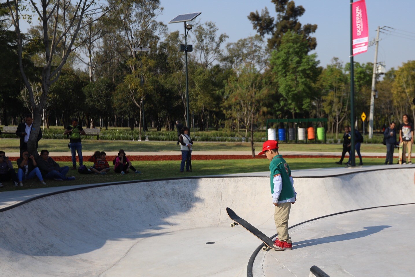 Ein junger Junge in einem grünen T-Shirt, braunen Hosen und einer roten Mütze fährt ein Skateboard die Seite einer Rampe auf einem Skatepark herunter, mit Menschen, Bäumen, Laternenpfählen und dem Himmel im Hintergrund.