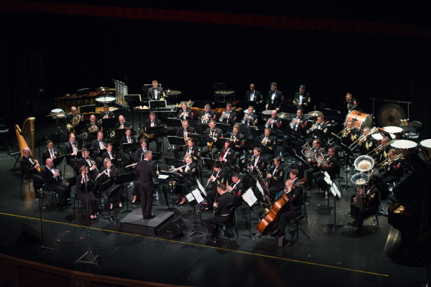 Ein großes Orchester auf einer Bühne mit einem Dirigenten in der Mitte, verschiedene Instrumente einschließlich Saxophone, Trompeten, Posaunen und Trommeln, sowie ein Dirigent in einem schwarzen Anzug und weißem Hemd, helle Bühnenbeleuchtung und ein Publikum im Hintergrund.