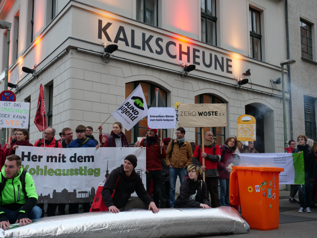 A group of people holding protest signs in front of a building in Germany, with two individuals seated in the foreground and a trash bin on the right.