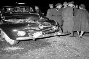 Ein Schwarz-Weiß-Bild eines auf der Straße liegenden, verbeulten Autos mit zersplitterten Fenstern, umgeben von einer besorgten Gruppe von Menschen.