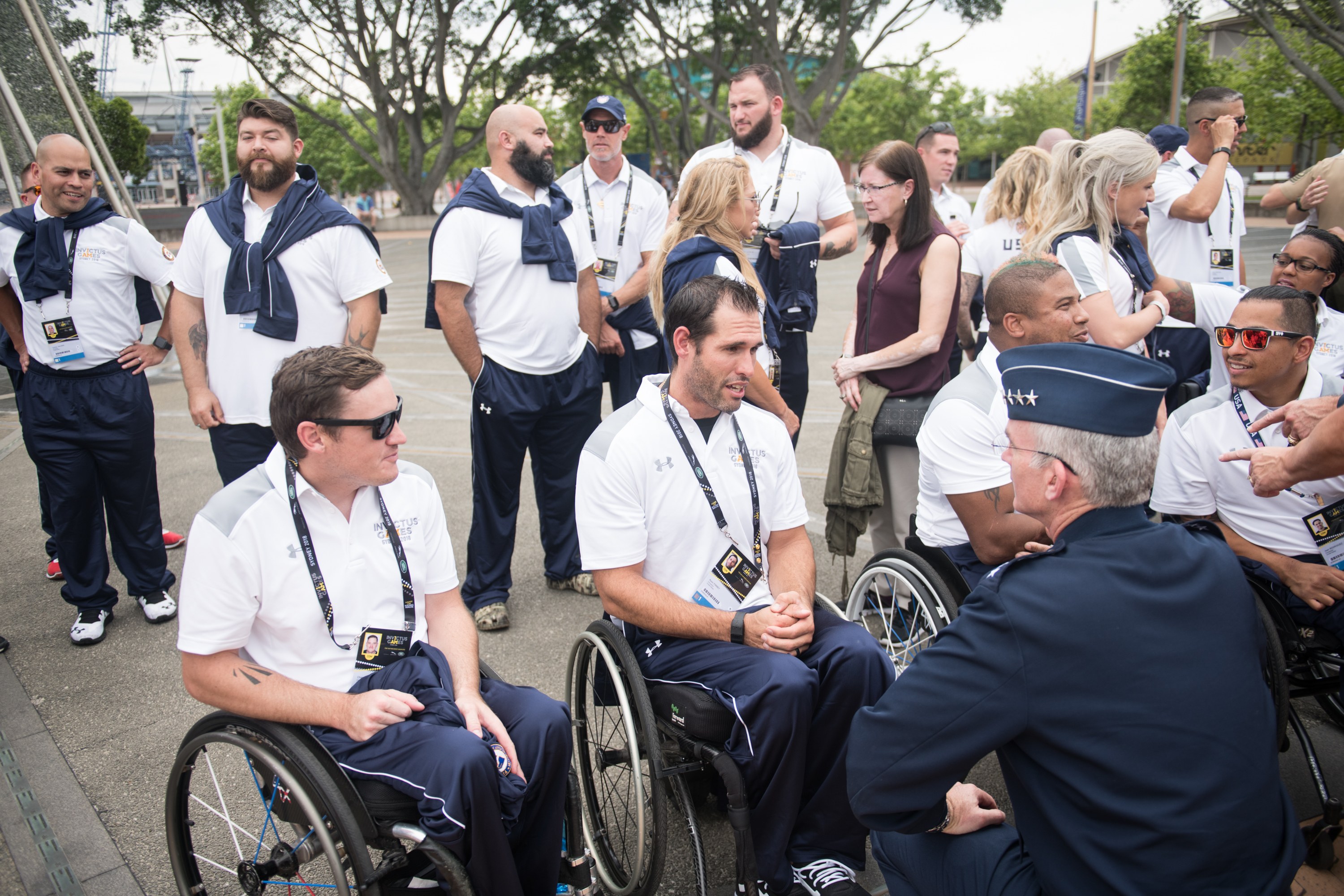 Eine Gruppe von Menschen in Rollstühlen vor einem Hintergrund aus Bäumen, Pfählen, Gebäuden und einem klaren blauen Himmel, einige tragen Mützen und Schutzbrillen.