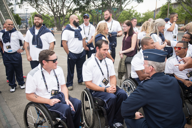 Eine Gruppe von Menschen in Rollstühlen vor einem Hintergrund aus Bäumen, Pfählen, Gebäuden und einem klaren blauen Himmel, einige tragen Mützen und Schutzbrillen.