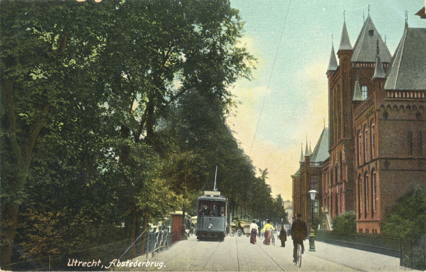 Altes Schwarz-Weiß-Foto einer Stadtstraße mit einer Tram, Gebäuden, Straßenlaternen, Lampen, Fahrzeugen, Fußgängern, Geländern, Bäumen und bewölktem Himmel; Text unten.