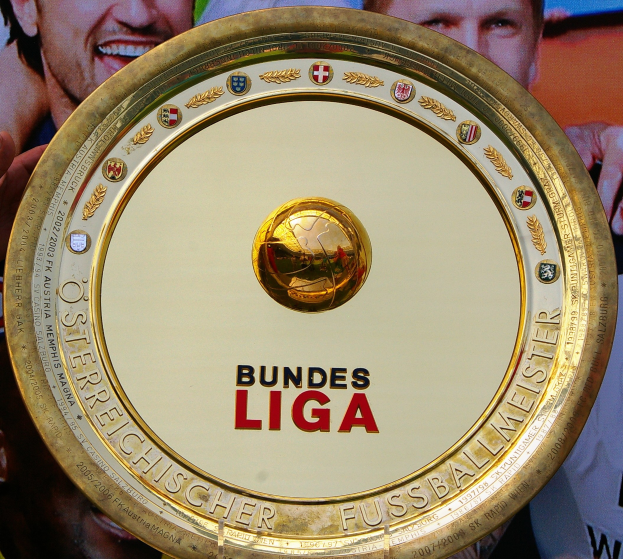 Ein Mann hält einen goldenen Schild mit der Aufschrift "Bundesliga", im Hintergrund ist eine Fahne mit drei Personen zu sehen.