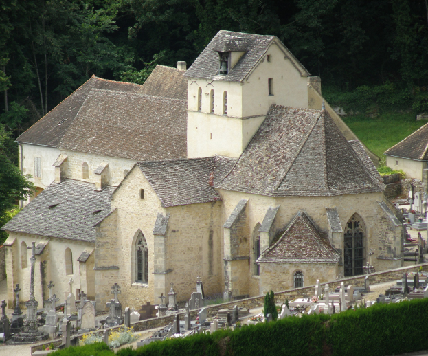 Alte Kirche mit einem Friedhof voller Grabsteine vorne, umgeben von Häusern, Bäumen, Pflanzen und Gras.