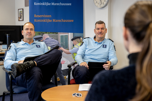 Zwei Polizeibeamte sitzen an einem Tisch mit Papieren und anderen Gegenständen und sprechen mit einer Frau. Im Hintergrund ist eine Wand mit einer Fahne und einer Uhr zu sehen. Links daneben steht ein weiterer Tisch mit einem Monitor, einer Tastatur, einer Maus und anderen Gegenständen.