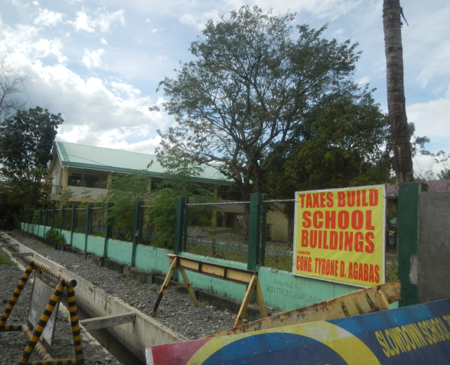 Eine Schule mit einem "Steuern Bauen Schulen"-Schild, umgeben von einem Zaun, Bäumen, Pflanzen, Gras, Steinen und einer Wand, unter einem bewölkten Himmel.