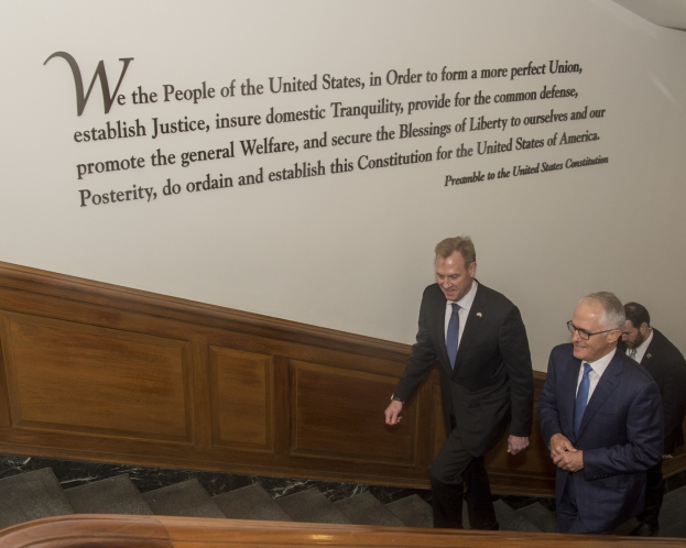 Two men in suits and ties walking down stairs, engaged in discussion, with one wearing glasses and a wall featuring text behind them.