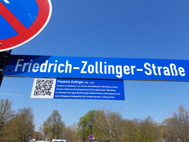 Straßenschild mit einem QR-Code, das vor einem Gebäude positioniert ist, mit Fahrzeugen auf der Straße, Bäumen und einem klaren blauen Himmel im Hintergrund.