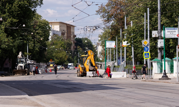 Stadtstraße mit einer Baustelle, Fahrzeugen, Fußgängern, einem Radfahrer, Verkehrsleitkegeln, Schildern, Versorgungsmasten mit Drähten, Bäumen, Gebäuden und einem bewölkten Himmel.