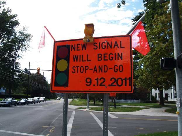 Straße mit Fahrzeugen, ein Verkehrszeichenmast mit einer "Neues Signal Beginn Stopp und Geh"-Plakette und ein Hintergrund aus Bäumen, Gebäuden und einem klaren blauen Himmel.