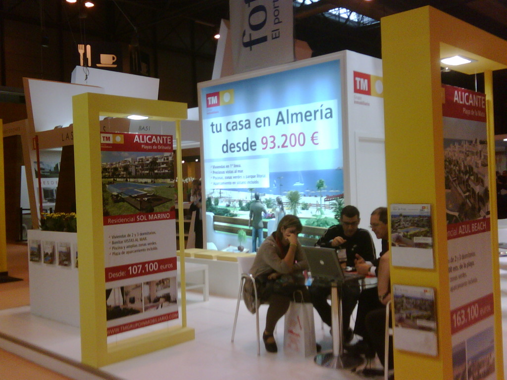 Eine Gruppe von Menschen sitzt um einen Tisch herum auf Stühlen und schaut auf einen Laptop, mit mehreren Flyern, auf denen verschiedene Werbeanzeigen zu sehen sind.