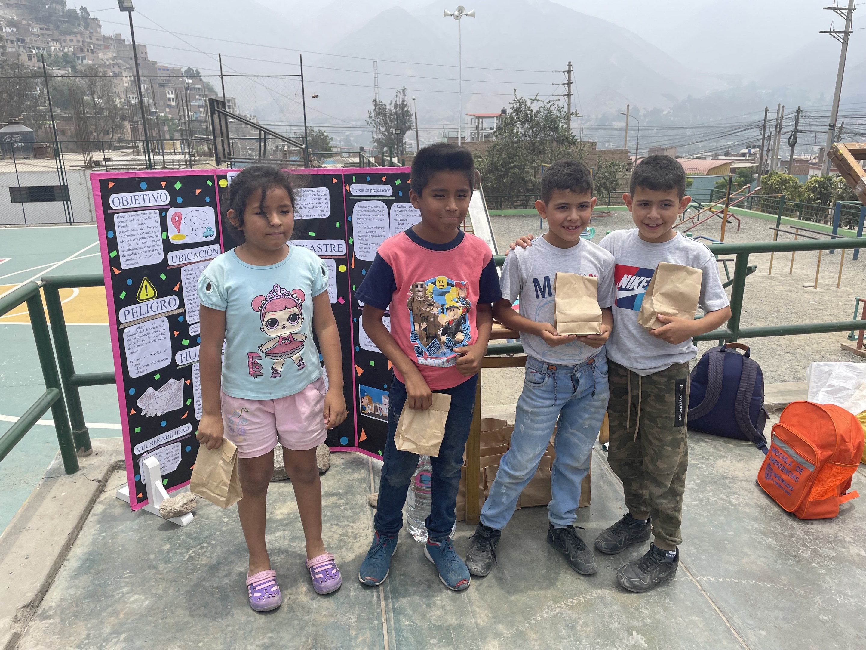 Gruppe von Kindern auf einem Basketballfeld mit Papiertüten, mit Vorräten auf dem Boden und einer Tafel mit Text links daneben, vor einer Outdoor-Bühne mit Zäunen, Pfosten und Gebäuden.