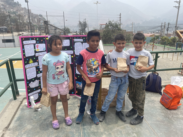 Gruppe von Kindern auf einem Basketballfeld mit Papiertüten, mit Vorräten auf dem Boden und einer Tafel mit Text links daneben, vor einer Outdoor-Bühne mit Zäunen, Pfosten und Gebäuden.