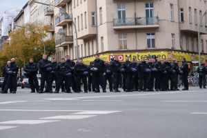 Eine Gruppe von Polizisten in schwarzen Uniformen mit blauen Helmen steht in der Mitte einer Straße, die von Laternenmasten, Bäumen und Gebäuden mit Glasfenstern und Balkonen gesäumt ist, mit weiteren Bäumen und einem klaren blauen Himmel im Hintergrund.