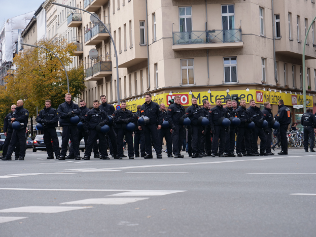 Eine Gruppe von Polizisten in schwarzen Uniformen mit blauen Helmen steht in der Mitte einer Straße, die von Laternenmasten, Bäumen und Gebäuden mit Glasfenstern und Balkonen gesäumt ist, mit weiteren Bäumen und einem klaren blauen Himmel im Hintergrund.