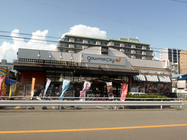 Eine belebte Stadtstraße mit hohen Gebäuden, Fahrzeugen, Straßenmöblierung, Grünflächen und einem Restaurant, das Speisen in seinem Schaufenster präsentiert.