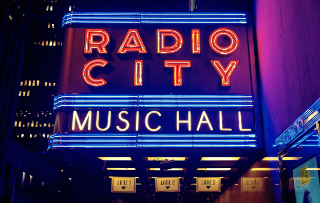 Hell erleuchtetes Radio City Music Hall in New York City mit hohen Gebäuden drumherum, mit einer 'Radio City'-Leuchtschrift und zusätzlichen Schildern rechts.