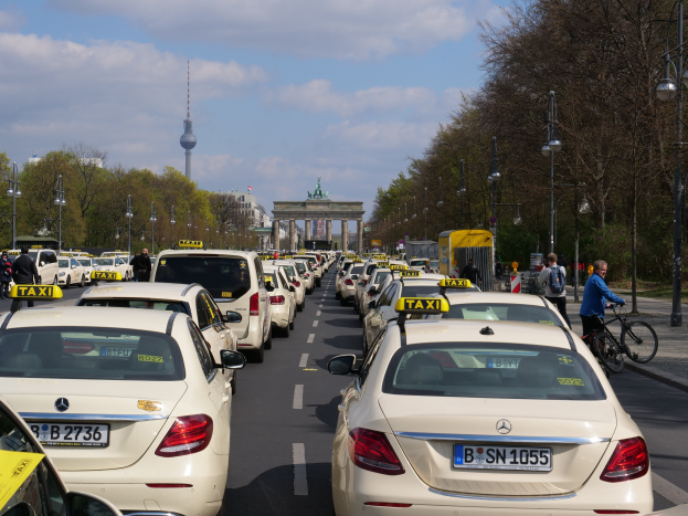 Eine lange Reihe gelber Taxis, die an einer belebten Straße in Berlin, Deutschland, geparkt sind, mit Fahrradfahrern und Fürstlichen auf dem Gehweg, flankiert von Bäumen und Laternenmästen und Gebäuden, einem Bogen und einem Turm im Hintergrund bei einem bewölktem Himmel.