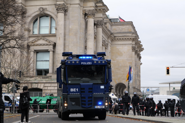 Gruppe von Polizeibeamten vor einem gro├čen Geb├Ąude mit Fenstern, Pfeilern und Bogeng├Ąngen, mit Fahrzeugen auf der Stra├če und einer Person mit einer Kamera auf der linken Seite.