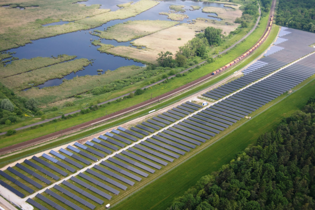 Luftaufnahme eines Solarparks mit Panelen, umgeben von Bäumen, Gras, Pflanzen und Wasser, mit einem Zug auf einer nahen Eisenbahnbrücke.