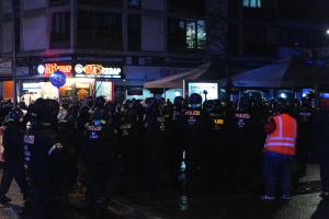 Eine Gruppe von Polizisten steht vor einer Menge auf einer Straße, umgeben von Gebäuden, Bäumen, Laternenmasten und Geschäften mit Schildern, mit einer Person mit Helm im Vordergrund.