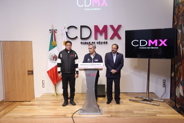 Drei Männer an einem Rednerpult während einer Pressekonferenz in Mexiko mit einem Mikrofon, Fernseher, Flaggen, einem Banner, einer Tür und einer Wand mit Text im Hintergrund.